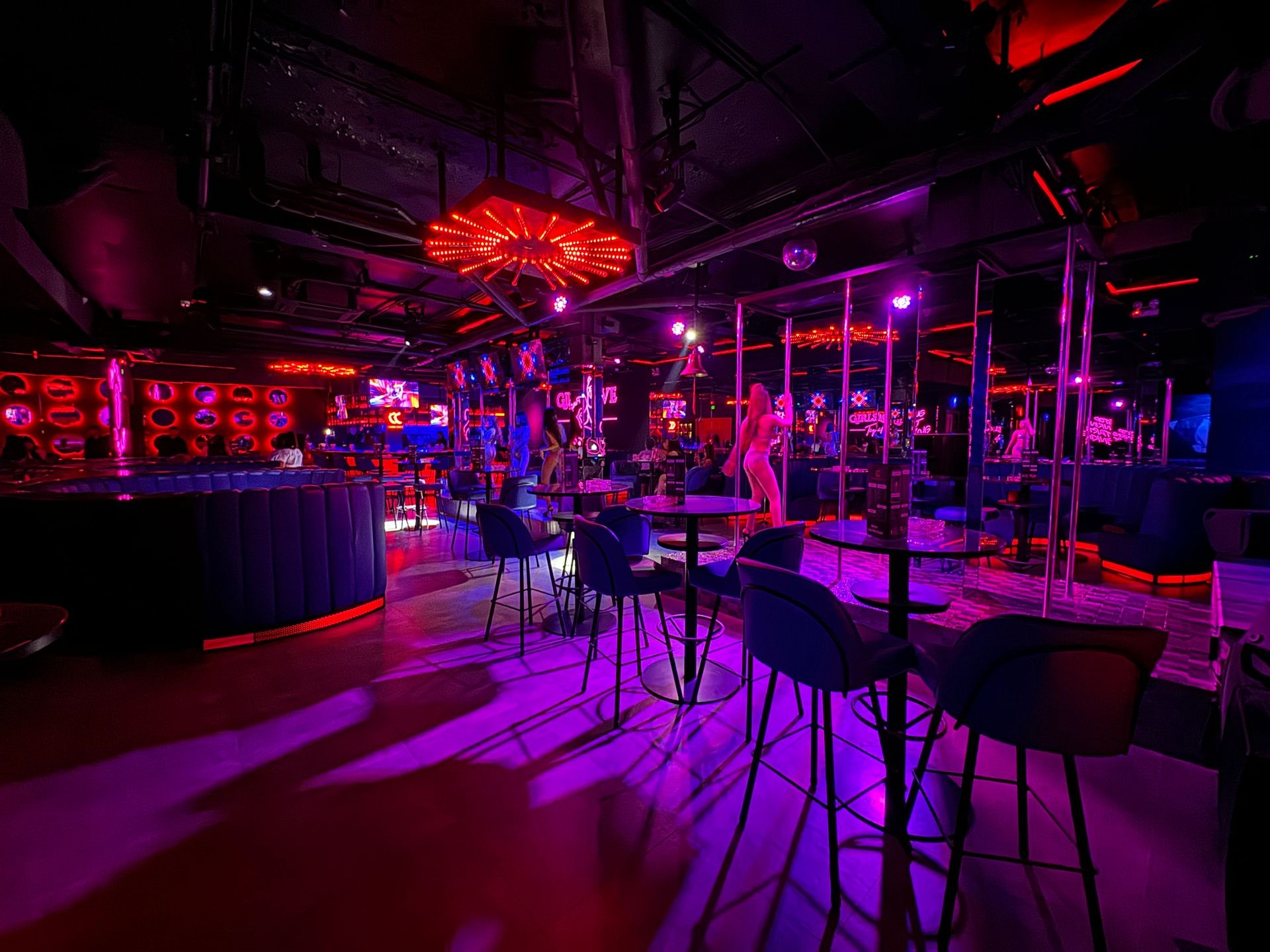 Dimly lit nightclub interior with neon lights, empty tables, and poles in the background.